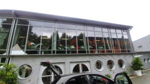 Modernes zweistöckiges Gebäude mit großen Glas- und Rundfenstern, Bäume spiegeln sich, Motorräder im Inneren, offener Kofferraum.