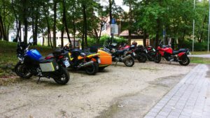 Motorräder, eines davon mit orangefarbenem Beiwagen, sind auf dem Schotter zwischen Bäumen und einem Gebäude geparkt.