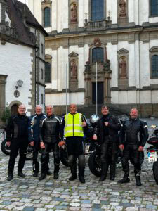 Sechs Biker in Ausrüstung posieren vor einem prächtigen historischen Gebäude; Motorräder reihen sich auf Kopfsteinpflaster auf.