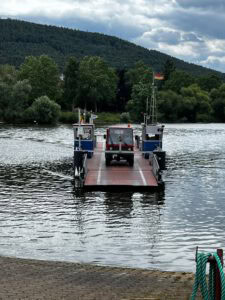 Eine kleine Fähre transportiert ein Nutzfahrzeug über einen ruhigen Fluss mit Hügeln, Bäumen und einer deutschen Flagge in der Nähe.