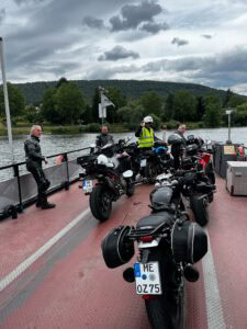 Vier Motorradfahrer in voller Montur stehen mit ihren Maschinen auf einer Fähre am Flussufer, dahinter üppige Hügel und wolkenverhangener Himmel.