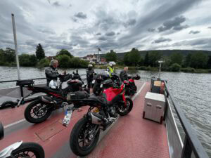 Vier Biker in voller Montur stehen mit ihren Motorrädern auf einer Fähre, hinter ihnen der Fluss und die Häuser.