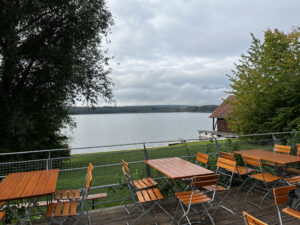 Leere Holztische und -stühle auf einer Terrasse mit Blick auf einen ruhigen See, umrahmt von Bäumen und einem kleinen Bootshaus.
