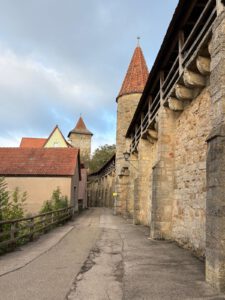Ein schmaler Pfad grenzt an eine mittelalterliche Steinmauer mit Holzstegen, rotgedeckten Türmen und Wolken darüber.