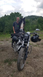 Ein Mann steht mit einem Pferd hinter einem geparkten Motorrad auf einem Feldweg, im Hintergrund Bäume und Hügel.