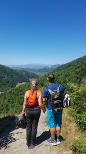 Ein händchenhaltendes Paar steht mit Helmen auf einem felsigen Pfad und bewundert ein üppiges Tal und die Berge.