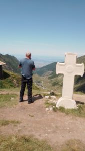 Ein Mann steht an einem weißen Kreuzdenkmal auf einem grasbewachsenen Bergrücken und überblickt eine kurvenreiche Straße und ein Tal.