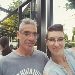 Ein lächelnder Mann und eine lächelnde Frau, die beide eine Brille tragen, machen ein Selfie in einem Café im Freien, das von Grün umgeben ist.