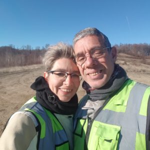 Ein lächelndes Paar mit Brille und Sicherheitsweste steht an einem sonnigen Tag mit trockenem Gras und Bäumen im Freien.