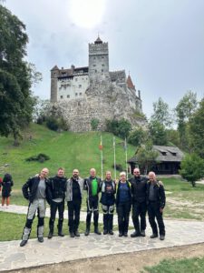 Acht Biker posieren vor dem Schloss Bran, mit Fahnenmasten, Grünpflanzen und einem rustikalen Holzhaus in der Nähe.