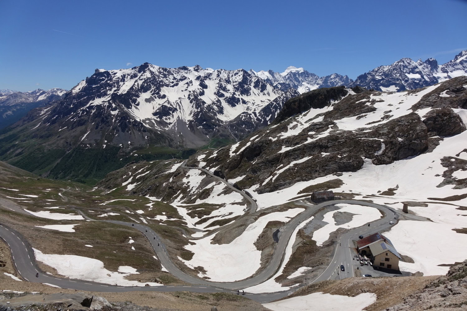 Eine kurvenreiche Bergstraße schlängelt sich durch den Schnee, vorbei an Berggipfeln, einigen Autos und einem Gebäude unter strahlend blauem Himmel.