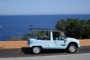Ein hellblaues Oldtimer-Cabrio steht vor einer orangefarbenen Mauer, mit Blick auf das Meer und Boote in der Ferne.
