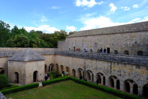 Ein historischer Steinarkadenhof umgibt einen begrünten Innenhof; Besucher schlendern unter einem teilweise bewölkten Himmel umher.
