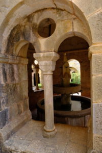Steinbögen und Säulen umrahmen einen sonnenbeschienenen Brunnen im Innenhof und bringen die verwitterten Steinstrukturen besonders gut zur Geltung.