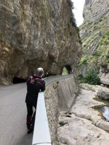 Ein Motorradfahrer in schwarz-roter Kleidung steht auf einer Bergstraße und blickt auf einen Tunnel über einem felsigen Fluss unter ihm.