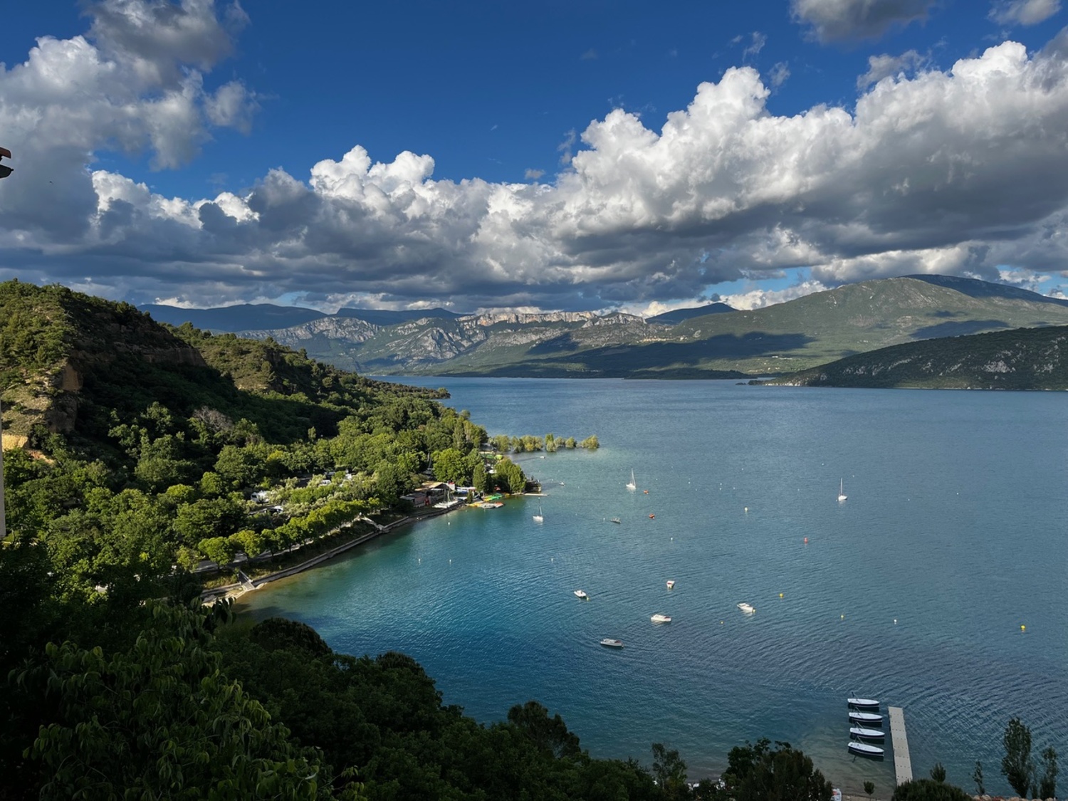 Ein ruhiger See, umgeben von üppig bewachsenen Hügeln und Bergen, auf dem hier und da Boote schwimmen und unter den Wolken ein Steg zu sehen ist.
