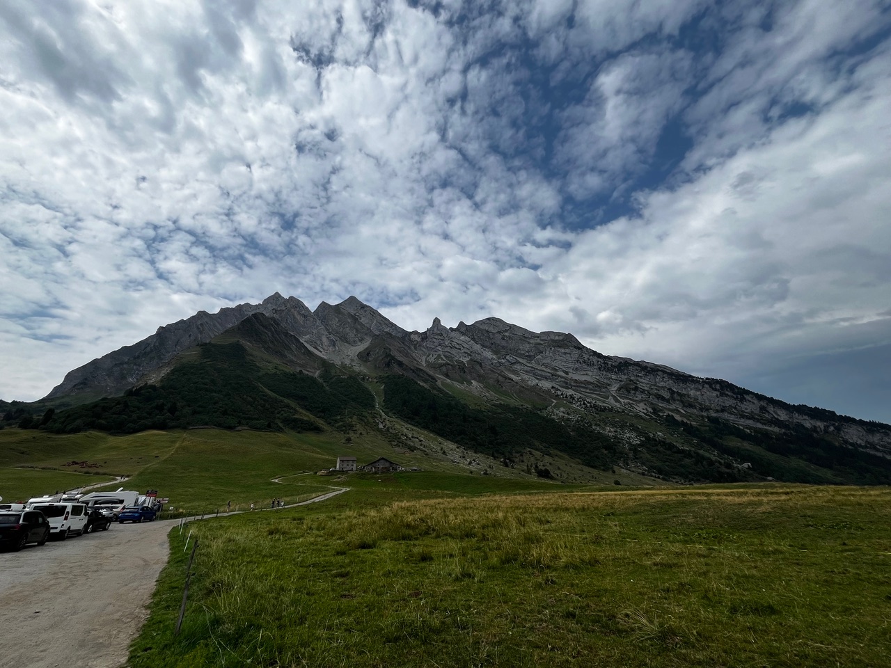 IMG_7485 Unter einem teilweise bewölkten Himmel ragen majestätische Berge empor und überragen grüne Wiesen und ein einsames Gebäude.