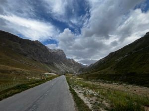 Eine schmale, asphaltierte Straße schlängelt sich durch ein üppig bewachsenes Bergtal unter einem dramatischen, wolkenverhangenen Himmel.