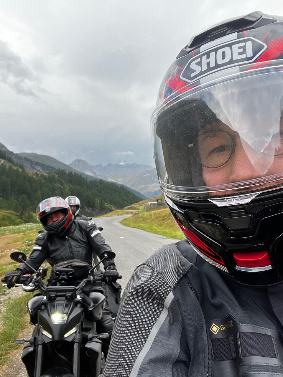 IMG_7502 Zwei Motorradfahrer in voller Montur lächeln, während sie eine kurvenreiche Bergstraße entlangfahren; einer von ihnen macht ein Selfie mit Helm.
