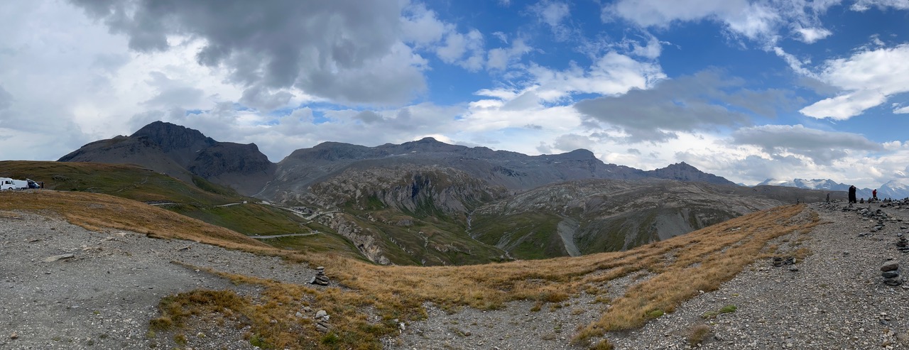 IMG_7508 Ein Panoramablick auf felsige Berge mit kurvenreichen Straßen, grasbewachsenen Hängen und einigen Menschen am Rand unter den Wolken.