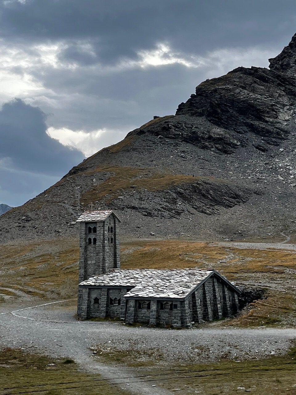 IMG_7511 Auf einem felsigen Hügel, unter bewölktem Himmel und neben einem gewundenen Feldweg steht eine kleine Steinkirche mit einem Glockenturm.