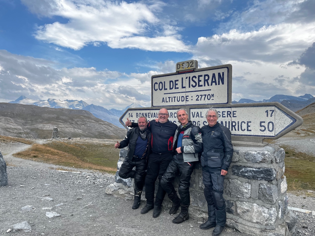 IMG_7518 Vier Männer in Motorradbekleidung lächeln vor einem Schild am Col de l’Iseran, im Hintergrund sind Berge und Wolken zu sehen.