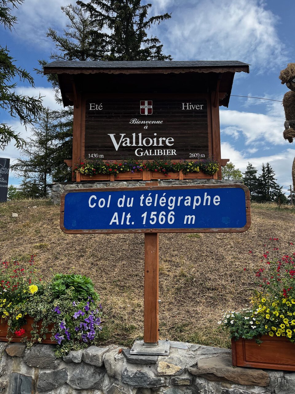 IMG_7528 Ein Holzschild mit der Aufschrift „Valloire Galibier“ und ein blaues Schild mit der Aufschrift „Col du Télégraphe, 1566 m“ inmitten von Blumen und Bäumen.