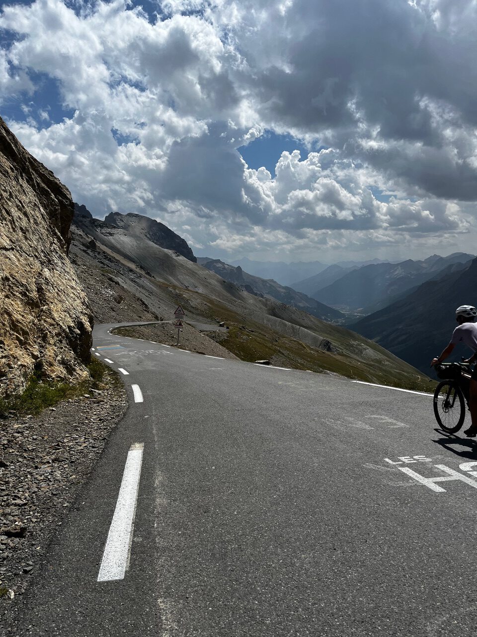 IMG_7539 Ein Radfahrer fährt eine kurvenreiche Bergstraße hinauf, während hinter ihm vereinzelte Wolken, schroffe Gipfel und Täler zu sehen sind.
