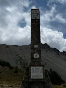 Am Col d’Izoard (2360 m) steht inmitten von felsigen Hügeln und wolkenverhangenen Gipfeln ein hohes Steinmonument mit Gedenktafeln.