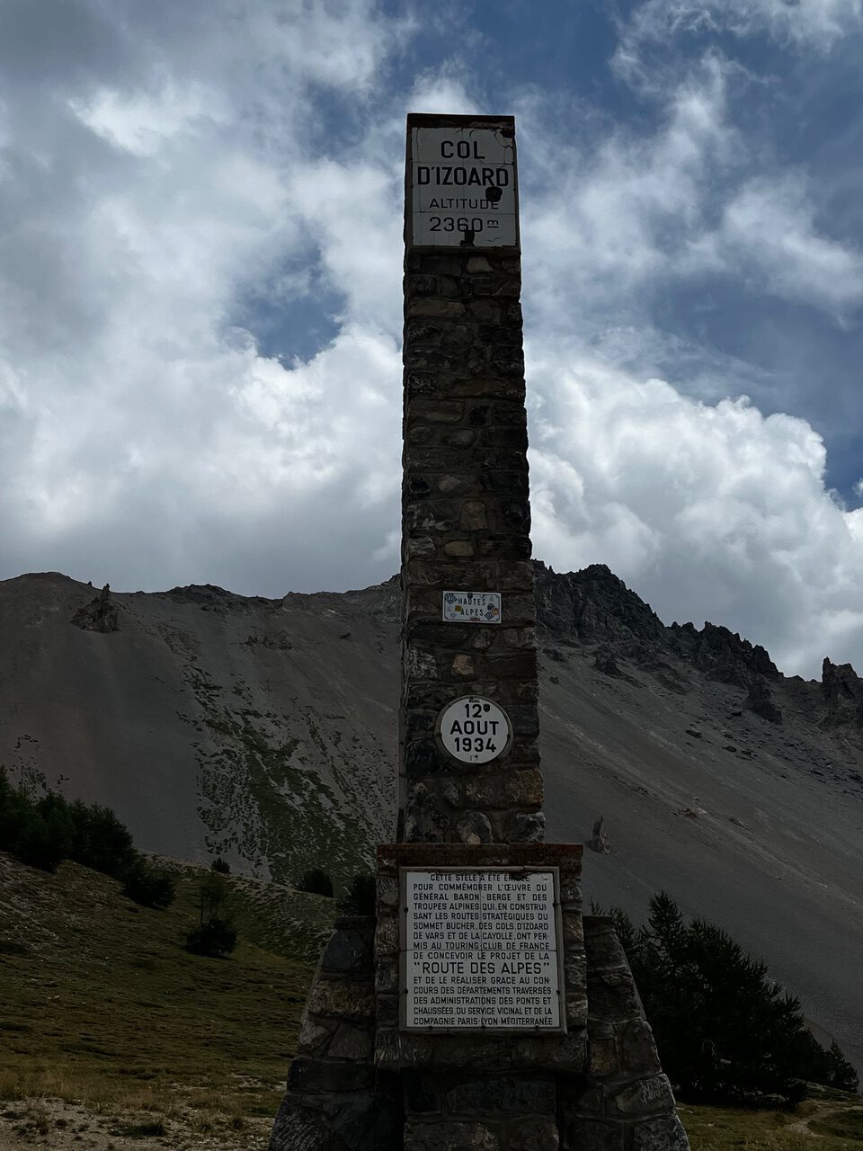 IMG_7550 Am Col d’Izoard (2360 m) steht inmitten von felsigen Hügeln und wolkenverhangenen Gipfeln ein hohes Steinmonument mit Gedenktafeln.