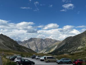 Ein Parkplatz in einem Bergtal mit Autos, felsigen Gipfeln, grüner Natur und einem blauen Himmel mit Wolken.