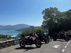Vier Motorräder, die unter blauem Himmel an einem Aussichtspunkt am See geparkt sind, im Hintergrund schattenspendende Bäume und Hügel.