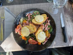 Salat mit Blattgemüse, Speckwürfeln, Tomatenschnitzen, gelben Tomaten und Ziegenkäse-Toast auf einem schwarzen Teller.