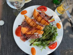 Mit Prosciutto umwickelte Cantaloupe-Melone mit Salat, Tomaten und Balsamico auf einem weißen Teller; daneben steht ein Glas Wein.