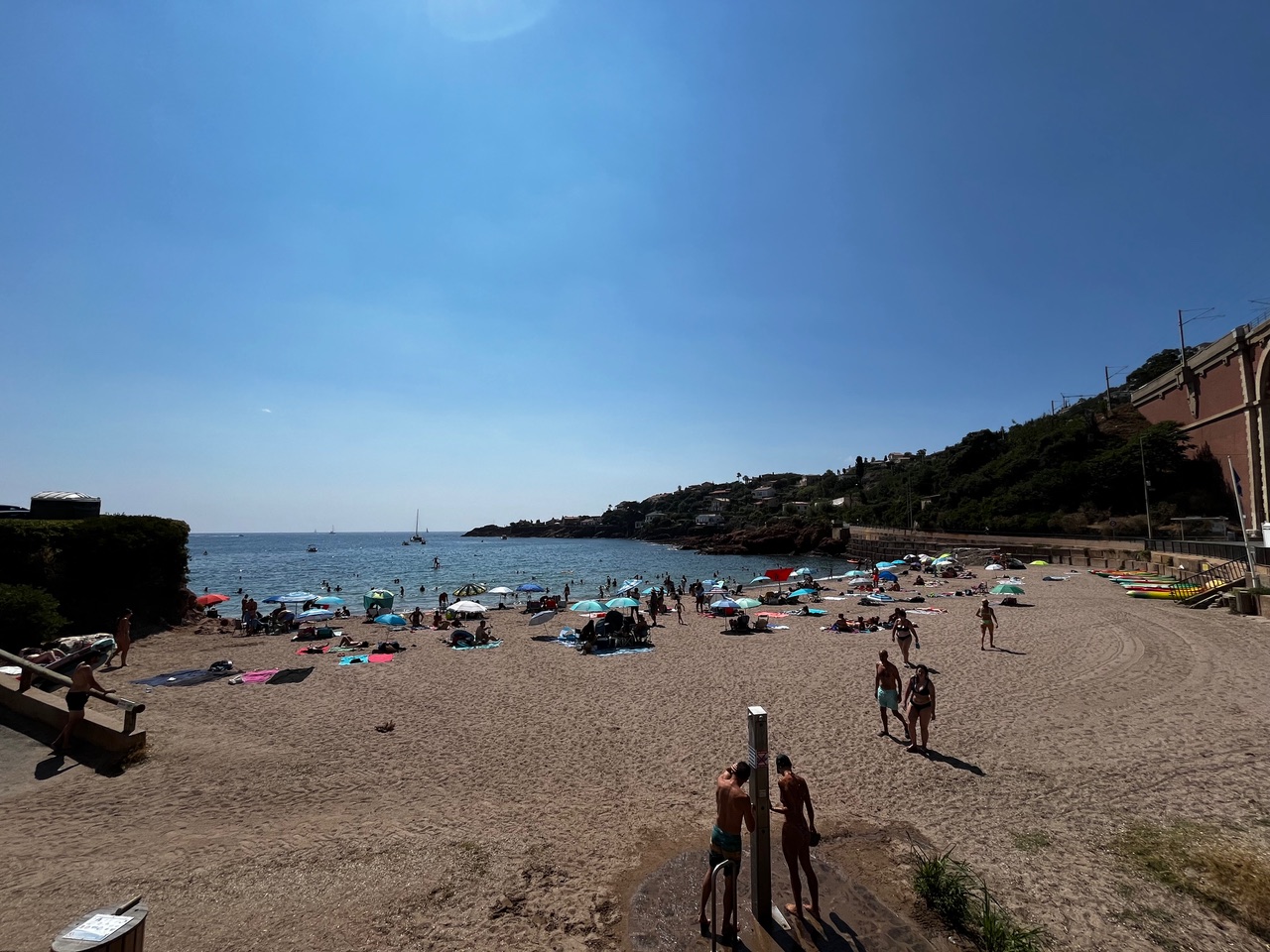 IMG_7607 An einem sonnigen Strand entspannen sich Menschen unter Sonnenschirmen; ruhiges Meer, Boote und Häuser am Hang in der Ferne.