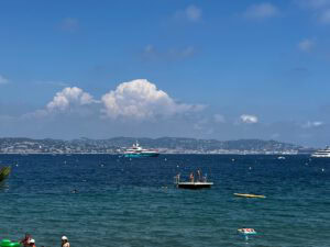 Menschen schwimmen an einem Schwimmsteg im blauen Meer vorbei, im Hintergrund sind vor Anker liegende Boote und die Skyline der Stadt zu sehen.