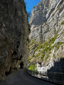 Eine schmale Straße schlängelt sich zwischen felsigen Klippen hindurch, mit einem Tunnel, einem Metallgeländer und einem strahlend blauen Himmel darüber.