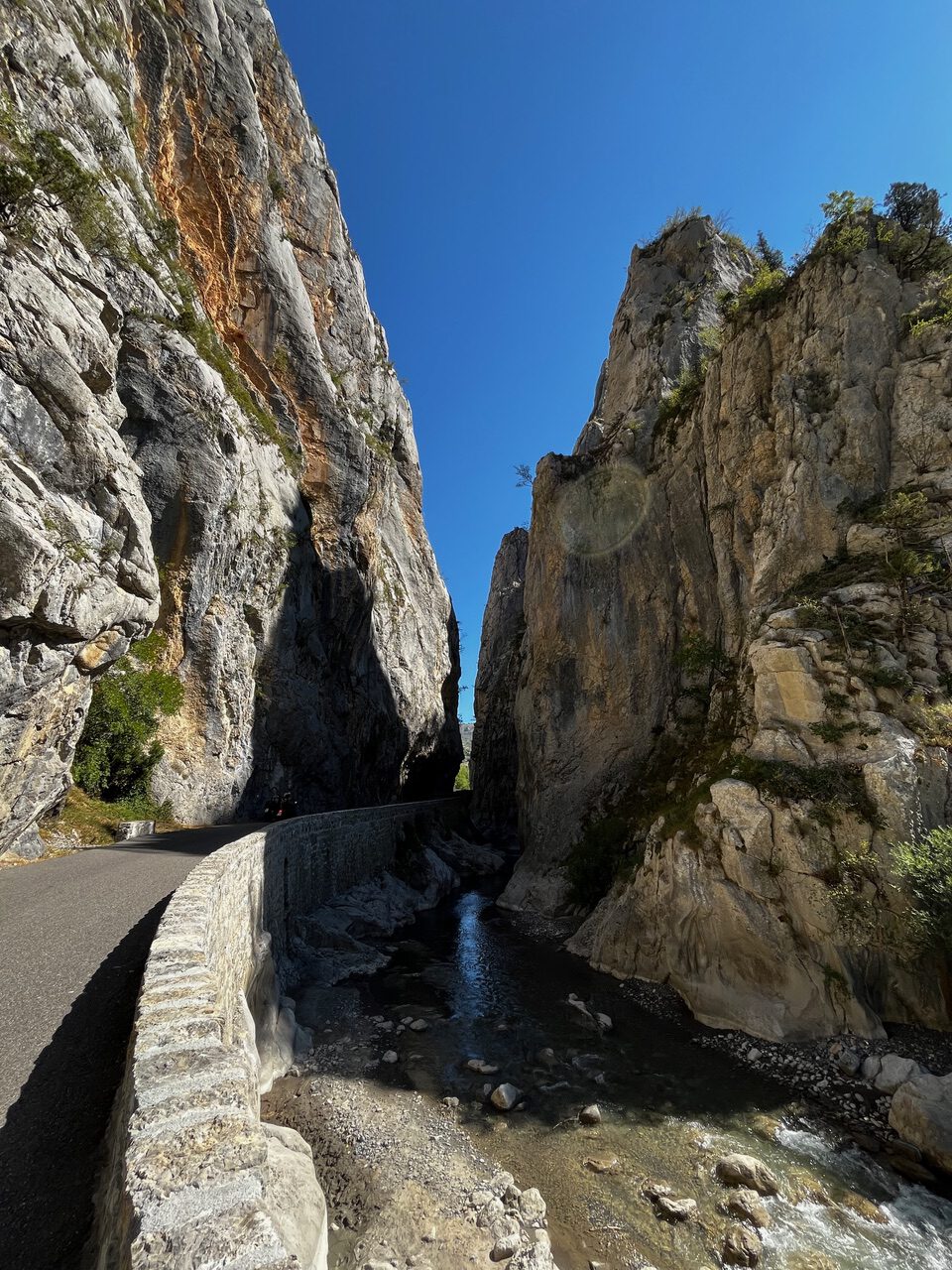 IMG_7649 Eine enge Bergschlucht mit steilen Felswänden, einer kurvenreichen Straße und einem seichten Bach unter blauem Himmel.