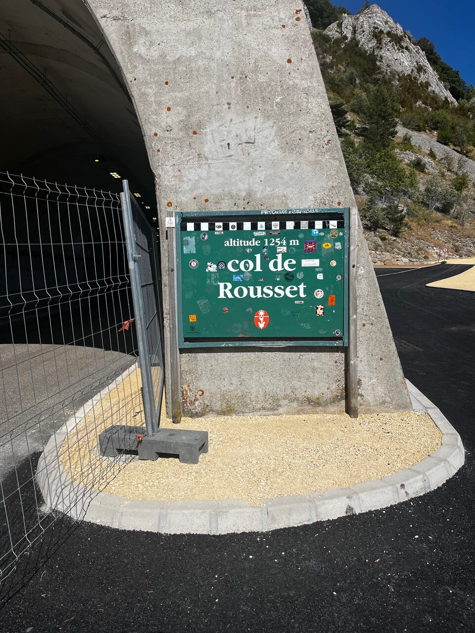 IMG_7658 Grünes Schild mit der Aufschrift „Höhe 1254 m, Col de Rousset“ auf Beton neben einem Tunnel, dahinter Berge.