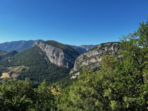 Unter einem blauen Himmel ragen schroffe Berge und felsige Klippen aus dichten Wäldern empor, eingerahmt von Ästen.