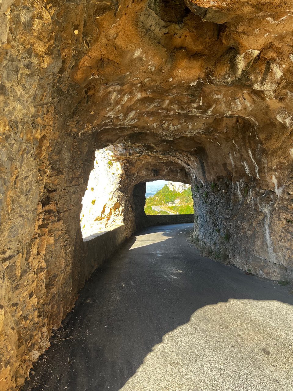 IMG_7673 Eine schmale Straße schlängelt sich durch einen felsigen Tunnel, während Sonnenlicht den Ausgang und den fernen Himmel dahinter erhellt.