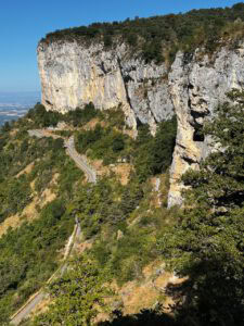 Eine kurvenreiche Bergstraße schlängelt sich unter steilen Felsen mit üppig grünen Bäumen hindurch, vor dem Hintergrund eines strahlend blauen Himmels.