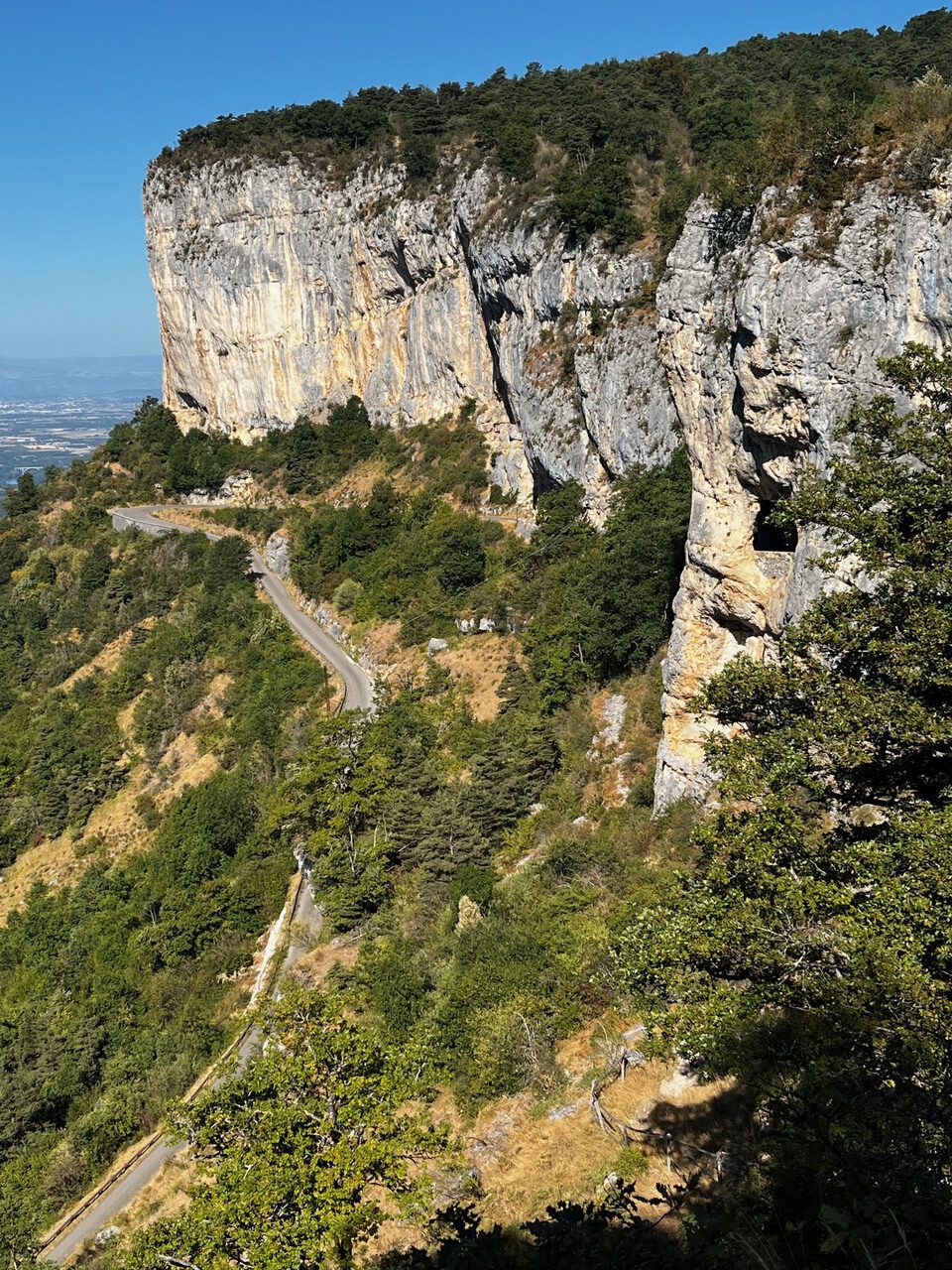 IMG_7676 Eine kurvenreiche Bergstraße schlängelt sich unter steilen Felsen mit üppig grünen Bäumen hindurch, vor dem Hintergrund eines strahlend blauen Himmels.