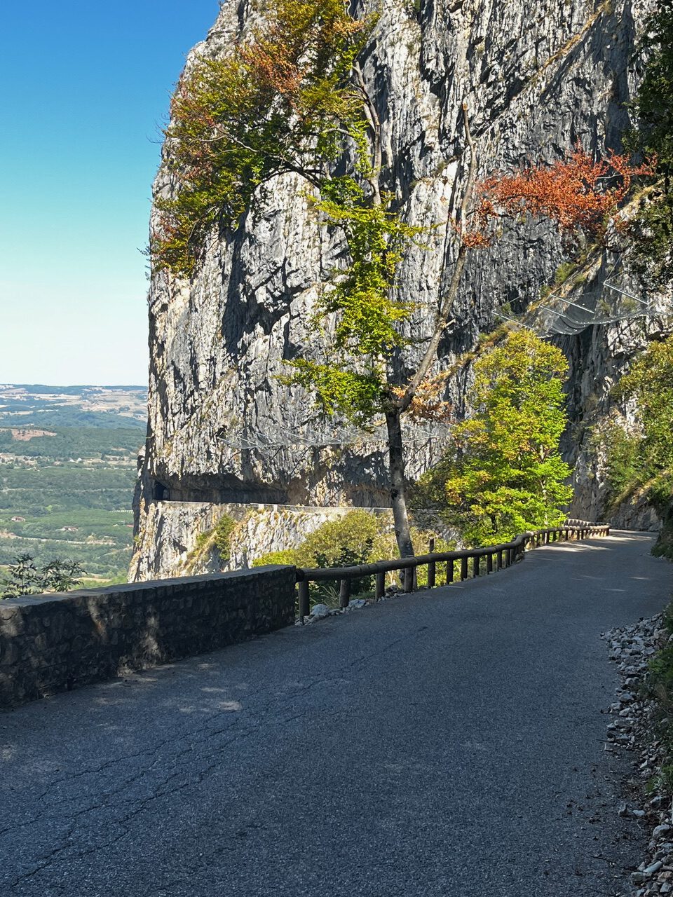IMG_7677 Eine kurvenreiche Bergstraße schlängelt sich an einer felsigen Klippe entlang, gesäumt von Bäumen und einem Geländer, mit Blick ins Tal.