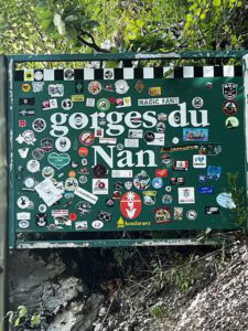 Ein grünes Schild mit der Aufschrift „Gorges du Nan“, das mit bunten Aufklebern beklebt ist, im Freien vor dem Hintergrund von Felsen und Laub.