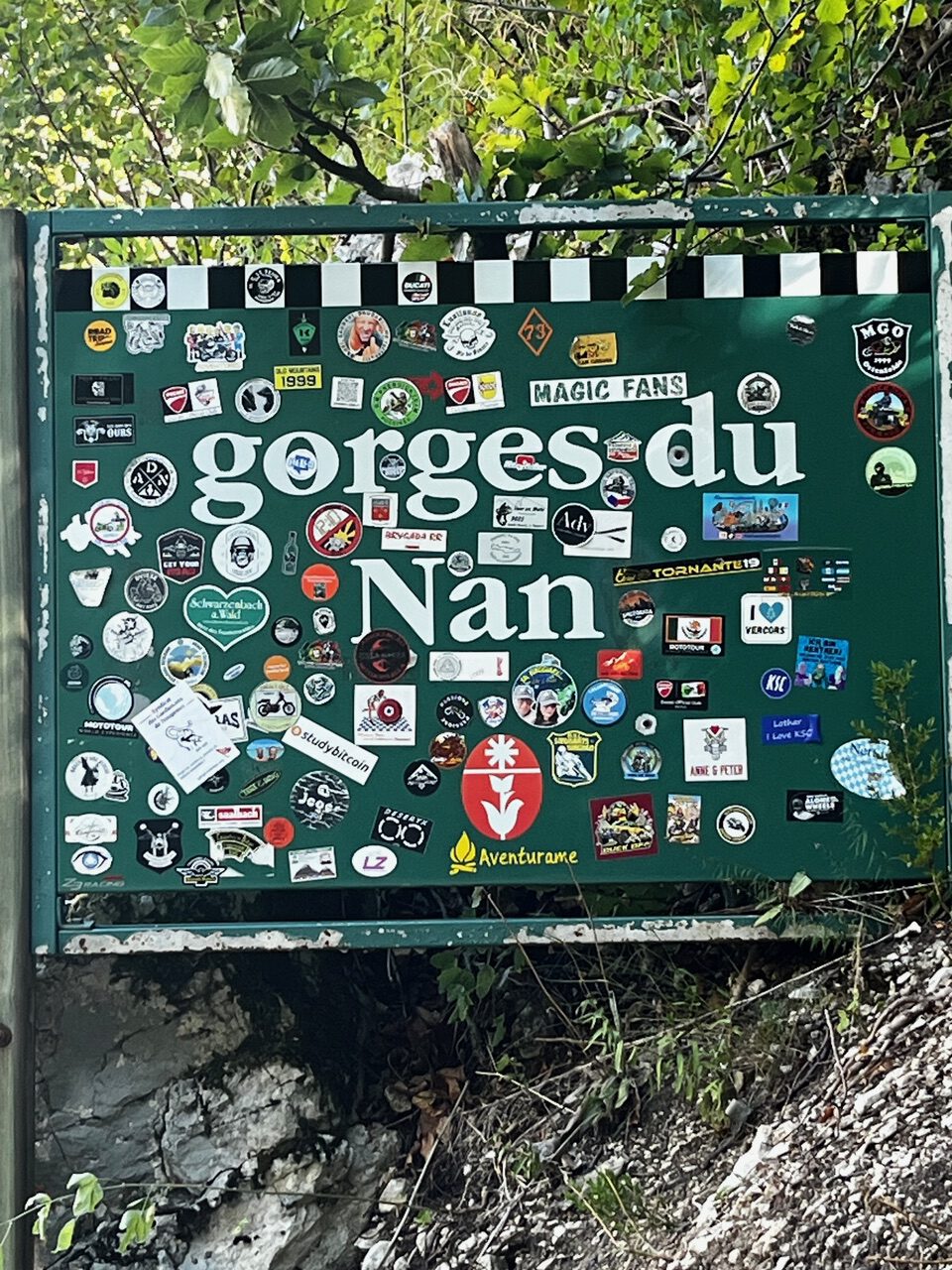 IMG_7678 Ein grünes „Gorges du Nan“-Schild im Freien, übersät mit Aufklebern, mit Felsen, Pflanzen und dem Aventurane-Logo.