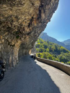 Eine kurvenreiche Bergstraße verläuft unter einem Felsvorsprung hindurch, gesäumt von Steinen und Grün; links ist ein Motorrad zu sehen.