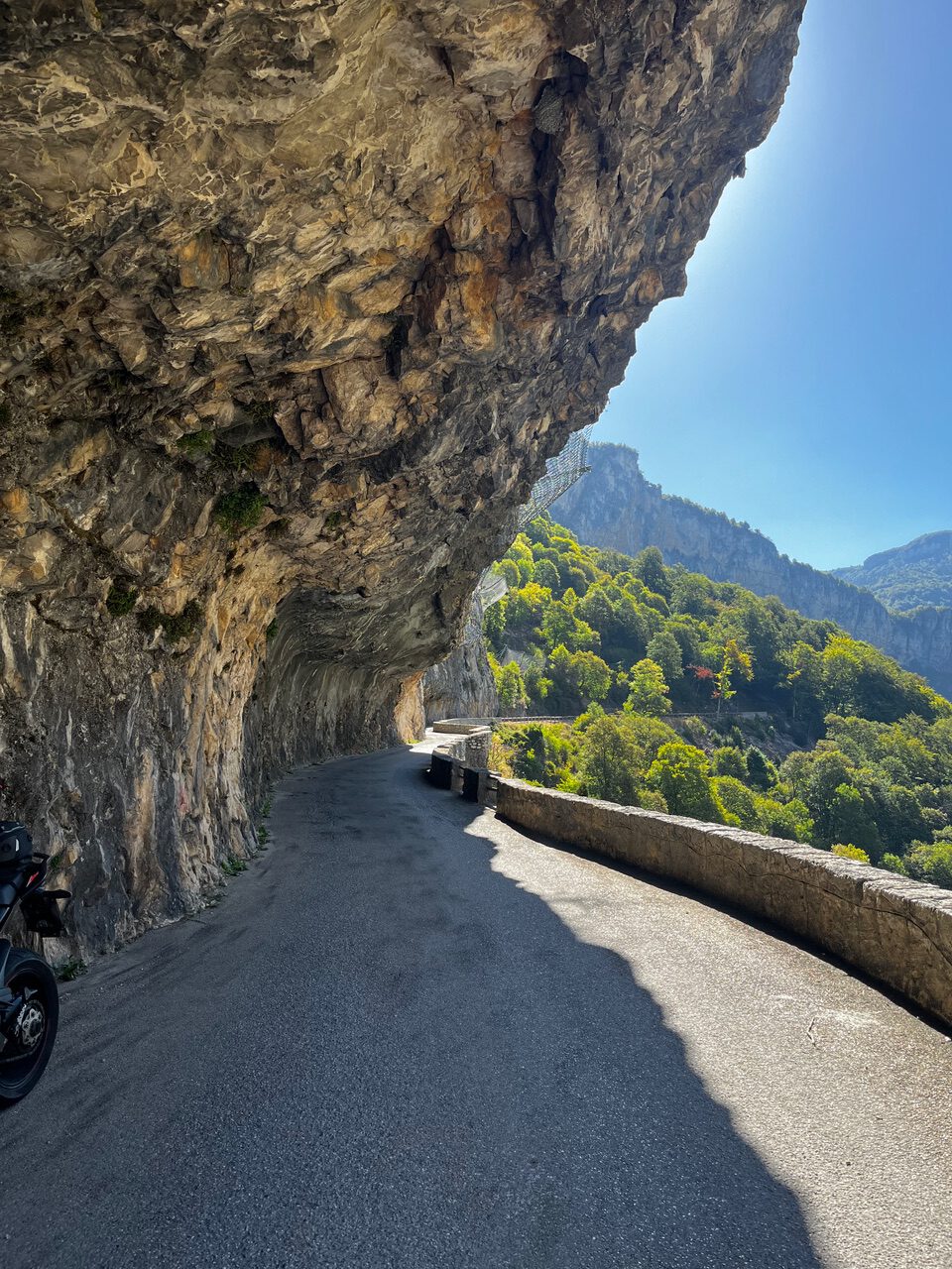 IMG_7679 Eine kurvenreiche Bergstraße verläuft unter einem Felsvorsprung hindurch, gesäumt von Steinen und Grün; links ist ein Motorrad zu sehen.