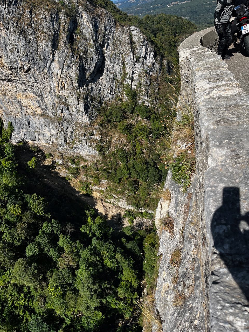 IMG_7680 Eine kurvenreiche Bergstraße schlängelt sich an einer steilen Felswand oberhalb eines dichten Waldes entlang; ein Motorradfahrer und sein Schatten fahren vorbei.