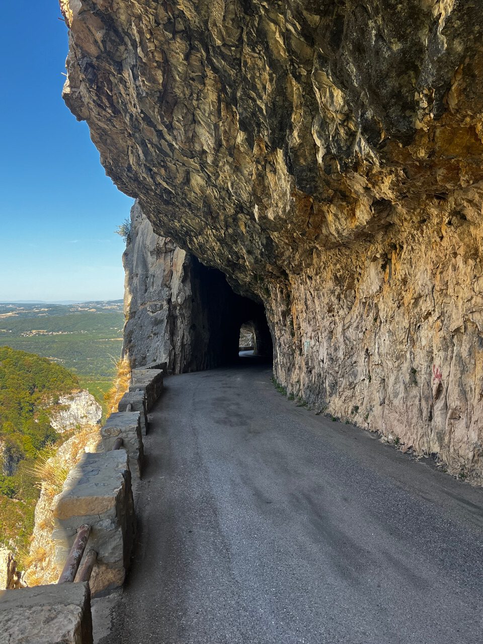 IMG_7681 Eine kurvenreiche Bergstraße führt durch einen Felsenstollen, an dessen Rändern Steinbarrieren stehen, dahinter erstreckt sich die Landschaft.